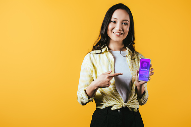 sonriente chica asiática apuntando a teléfono inteligente con aplicación de compras, aislado en amarillo
 - Foto, Imagen