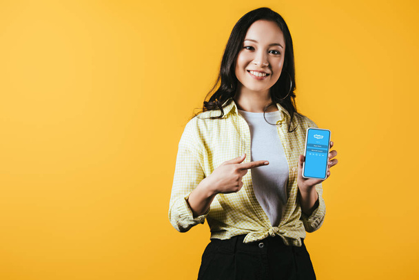 KYIV, UKRAINE - APRIL 16, 2019: attractive smiling asian girl pointing at smartphone with skype app, isolated on yellow - Foto, afbeelding
