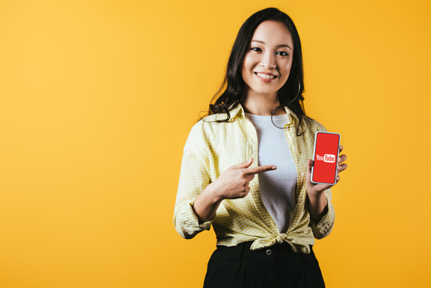 KYIV, UCRANIA - 16 DE ABRIL DE 2019: atractiva chica asiática sonriente apuntando al teléfono inteligente con la aplicación de youtube, aislada en amarillo
 - Foto, imagen