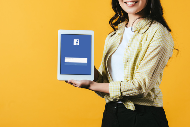 KYIV, UKRAINE - APRIL 16, 2019: cropped view of smiling girl showing digital tablet with facebook app, isolated on yellow - Photo, image
