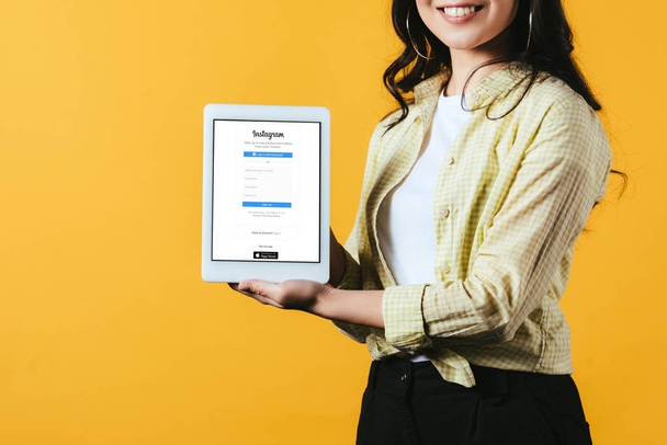 KYIV, UCRANIA - 16 DE ABRIL DE 2019: vista recortada de una chica sonriente mostrando una tableta digital con aplicación instagram, aislada en amarillo
 - Foto, Imagen