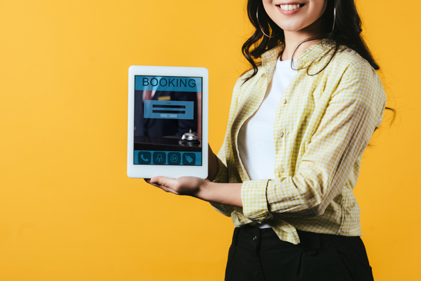 обрезанный вид девушки, показывающей цифровой планшет с приложением бронирования, изолированный на желтый
 - Фото, изображение