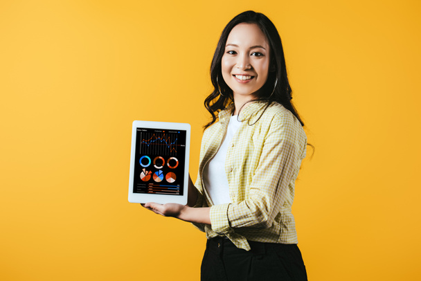 счастливая азиатская девушка показывает цифровой планшет с инфографикой, изолированный на желтый
 - Фото, изображение