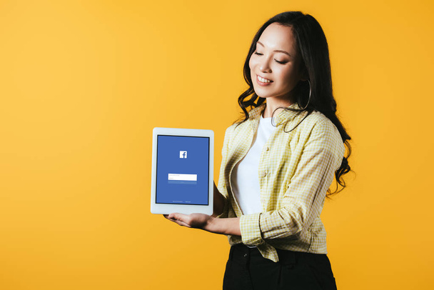 KYIV, UKRAINE - APRIL 16, 2019: beautiful asian girl showing digital tablet with facebook app, isolated on yellow - Foto, Imagem