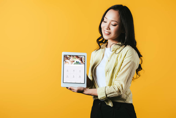 КИЕВ, УКРАИНА - 16 апреля 2019 года: красивая азиатская девушка показывает цифровой планшет с четырехквадратным приложением, изолированным на желтом
 - Фото, изображение