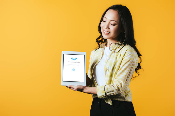 KYIV, UKRAINE - APRIL 16, 2019: beautiful asian girl showing digital tablet with skype app, isolated on yellow - Фото, зображення