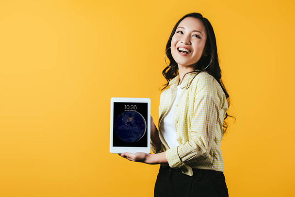 KYIV, UKRAINE - APRIL 16, 2019: smiling asian girl showing digital tablet screen, isolated on yellow - Fotó, kép