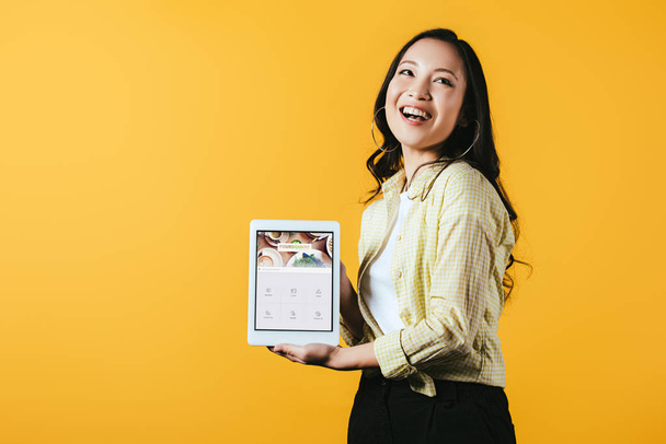 КИЕВ, УКРАИНА - 16 апреля 2019 года: красивая азиатская девушка показывает цифровой планшет с четырехквадратным приложением, изолированным на желтом
 - Фото, изображение