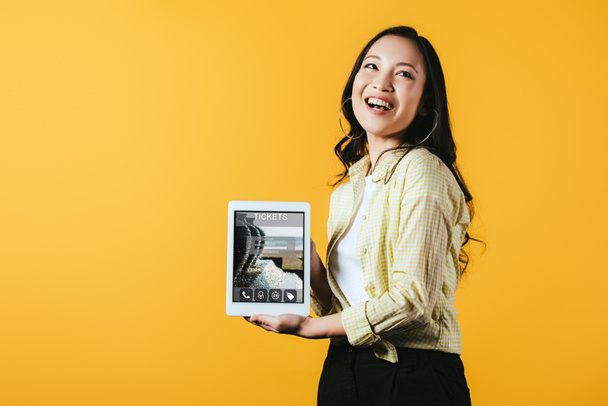 smiling asian girl showing digital tablet with tickets app, isolated on yellow - Photo, Image