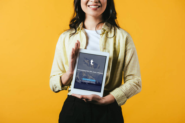 KYIV, UCRANIA - 16 DE ABRIL DE 2019: vista recortada de una chica sonriente mostrando una tableta digital con aplicación tumblr, aislada en amarillo
 - Foto, Imagen