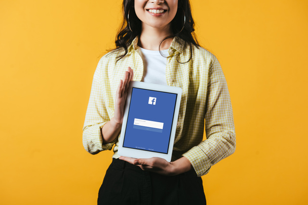 KYIV, UKRAINE - APRIL 16, 2019: cropped view of smiling girl showing digital tablet with facebook app, isolated on yellow - Photo, image