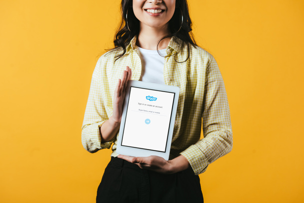 KYIV, UCRANIA - 16 DE ABRIL DE 2019: vista recortada de una chica sonriente mostrando una tableta digital con aplicación skype, aislada en amarillo
 - Foto, Imagen