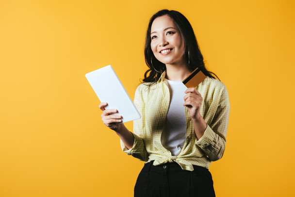 verträumte asiatische Mädchen online einkaufen mit digitalem Tablet und Kreditkarte, isoliert auf gelb - Foto, Bild