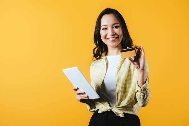 glimlachend Aziatisch meisje online winkelen met digitale Tablet en creditcard, geïsoleerd op geel - Foto, afbeelding