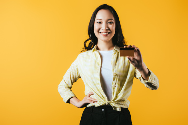 sonriendo chica asiática sosteniendo tarjeta de crédito, aislado en amarillo
 - Foto, Imagen