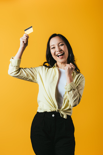 взволнованная азиатская девушка держит кредитную карту, изолированные на желтый
 - Фото, изображение