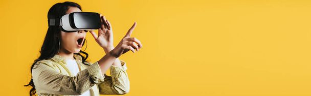 podekscytowany dziewczyna wskazując i korzystanie z zestawu słuchawkowego wirtualnej rzeczywistości, izolowane na żółtym  - Zdjęcie, obraz