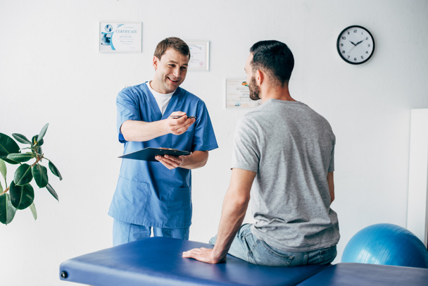 smiling Physiotherapist with diagnosis and pen gesturing near patient in hospital - Photo, Image