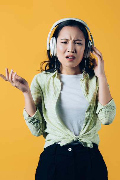 zmatené asijské dívky poslouchající hudbu se sluchátky, izolované na žluté - Fotografie, Obrázek