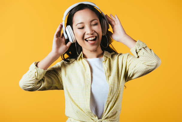 attractive asian girl singing and listening music with headphones, isolated on yellow - Foto, Imagen