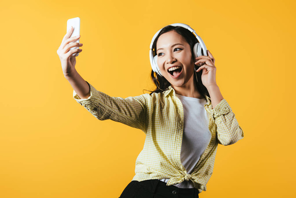 attraktive asiatische Mädchen Selfie auf dem Smartphone, singen und Musik hören mit Kopfhörern, isoliert auf gelb - Foto, Bild