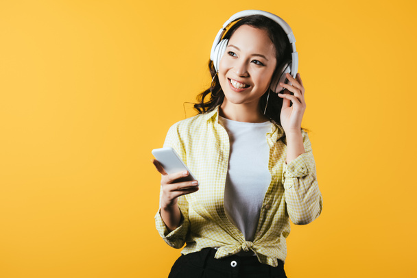 smiling asian girl listening music with headphones and smartphone, isolated on yellow - Foto, Imagen