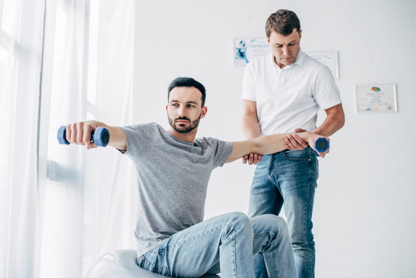 chiropractor stretching arm of good-looking patient with dumbbells in hospital - Photo, image