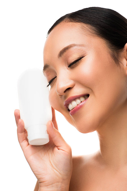 atrakcyjna, uśmiechnięta Azjatycka kobieta z zamkniętymi oczami trzymając butelkę balsamu, wyizolowana na białym tle - Zdjęcie, obraz