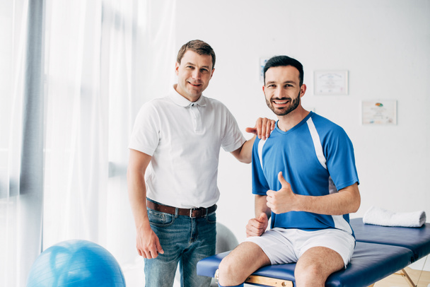 χαμογελαστός φυσιοθεραπευτής κοντά σε ευτυχισμένο Ποδοσφαιρικό παίκτη που δείχνει τον αντίχειρά του στο νοσοκομείο - Φωτογραφία, εικόνα
