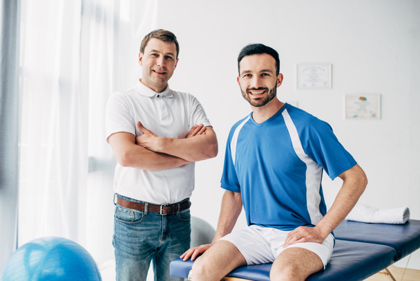 sonriente fisioterapeuta y jugador de fútbol en el hospital mirando a la cámara
 - Foto, Imagen