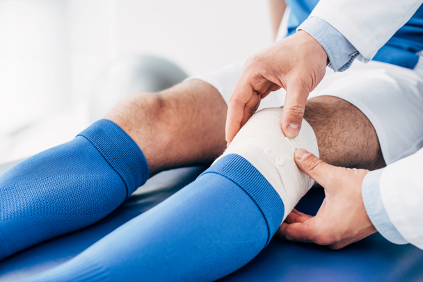 partial view of Physiotherapist putting elastic bandage on leg of football player - Valokuva, kuva