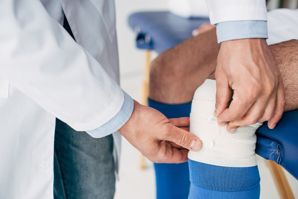 partial view of Physiotherapist in white coat putting elastic bandage on leg of football player - Φωτογραφία, εικόνα