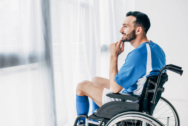 smiling man in football uniform sitting in Wheelchair and looking through window - Фото, изображение