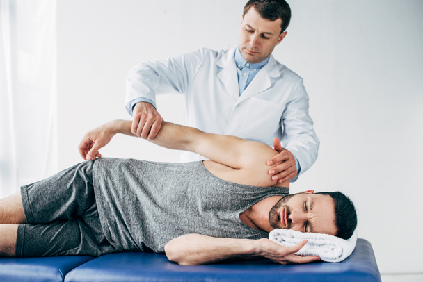 chiropractor stretching arm of handsome patient lying on massage table in hospital - Zdjęcie, obraz