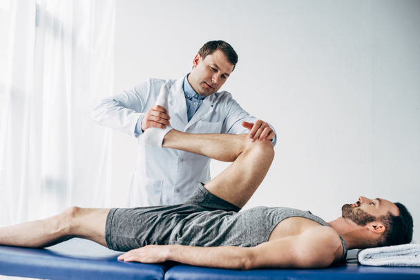 handsome chiropractor stretching leg of patient in hospital - Photo, Image