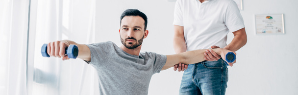 panoramic shot of chiropractor stretching arm of handsome patient with dumbbells in hospital - Photo, Image