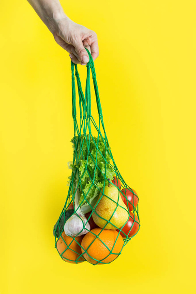 Mesh bag of fruits, vegetables, greens in woman's hand. Zero waste. Plastic free. Yellow background. - Fotó, kép