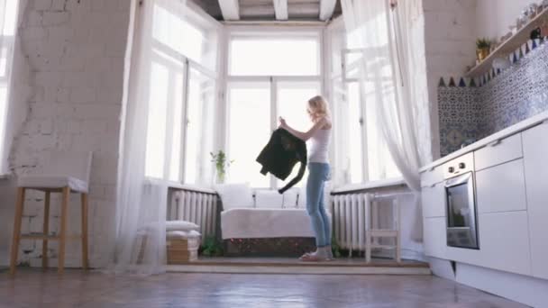 Close-up of beautiful blond woman in white t-shirt and jeans standing near the window and putting on a black jumper in her appartments. Action Relaxing at home - Filmati, video