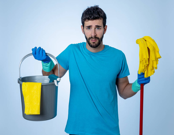 Young attractive man tired and upset holding bucket mop and cleaning spray feeling sad frustrated and lazy in domestic duties concept and men and women gender roles. Isolated on blue background - Fotografie, Obrázek