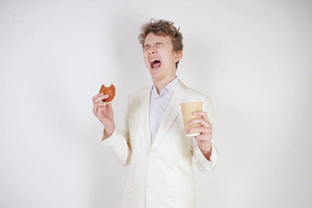 mladý běloch v bílém obleku stojí s papírovým šálkem kávy a buchtou v ruce a trpí těžkou bolestí břicha - Fotografie, Obrázek