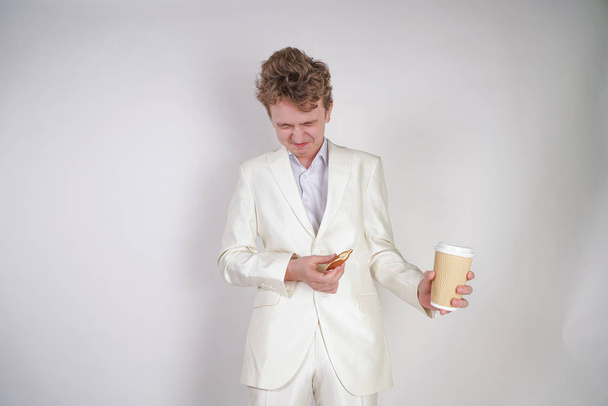Молодой кавказский мужчина в белом деловом костюме стоит с бумажной чашкой кофе и булочкой в руке и страдает от сильной боли в животе
 - Фото, изображение