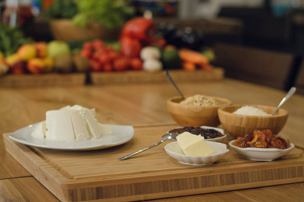 Desayuno diario con mantequilla, queso y mermelada sobre una mesa de madera. Hortalizas y frutas en el fondo
 - Foto, Imagen
