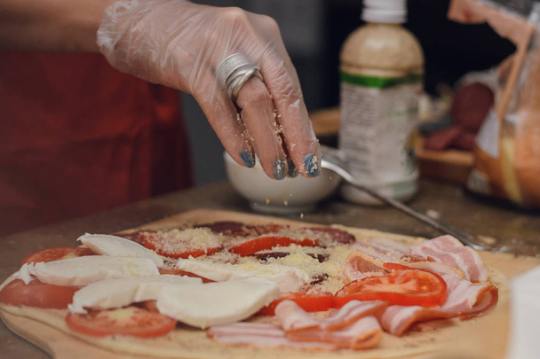 Een vrouw in wegwerphandschoenen hagelslag kaas pizza met ham en tomaten. Pizza koken - Foto, afbeelding