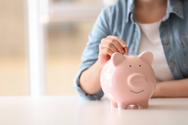 Woman putting coin into piggy bank at table indoors, closeup. Space for text - Foto, Imagen