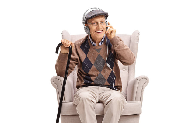 Elderly man sitting in an armchair and listening to music on earphones isolated on white background - 写真・画像