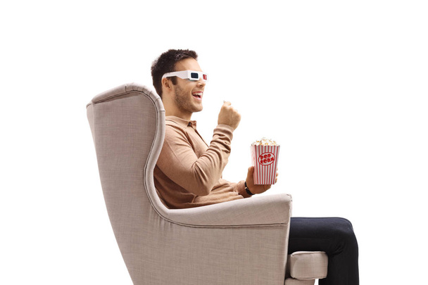 Young man in an armchair wearing a pair of 3D glasses and eating popcorn isolated on white background - Fotó, kép