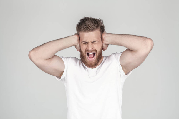 Portrait of beautiful sexy bearded man. holds his arms behind his head and shouts. he has a headache, he stands in front of the white background - Фото, зображення