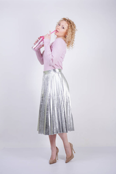 beautiful happy caucasian woman with curly blonde hair stands in a thin pink knitted sweater and a metallic silver pleated long skirt with a drink in her hands on a white background in the Studio - Photo, Image