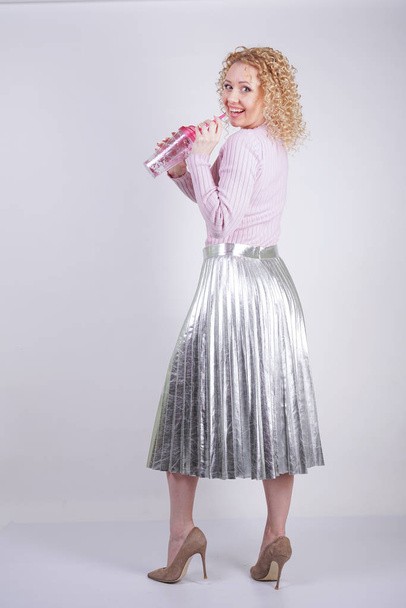 beautiful happy caucasian woman with curly blonde hair stands in a thin pink knitted sweater and a metallic silver pleated long skirt with a drink in her hands on a white background in the Studio - Foto, Bild