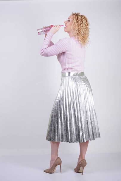 piękna szczęśliwa kaukaski kobieta z Kręcone blond włosy stoi w Cienki różowy sweter z dzianiny i metaliczny srebrny plisami długą spódnicę z drinkiem w dłoniach na białym tle w Studio - Zdjęcie, obraz